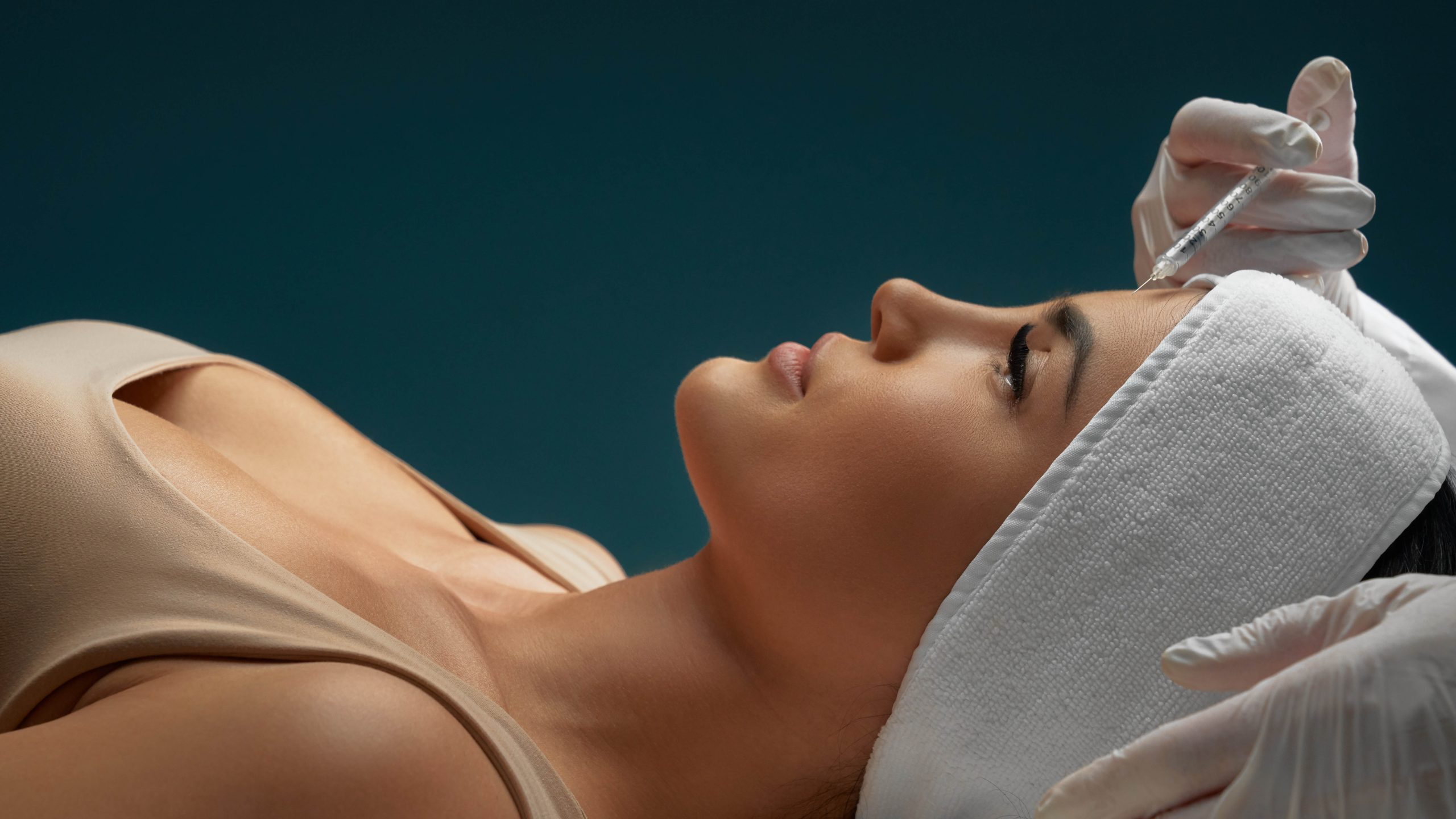 Close up of cosmetic botox injection in female forehead. Cosmetologist using syringe with liquid while brunette patient in towel on head lying, isolated on dark blue. Concept of cosmetology, beauty.